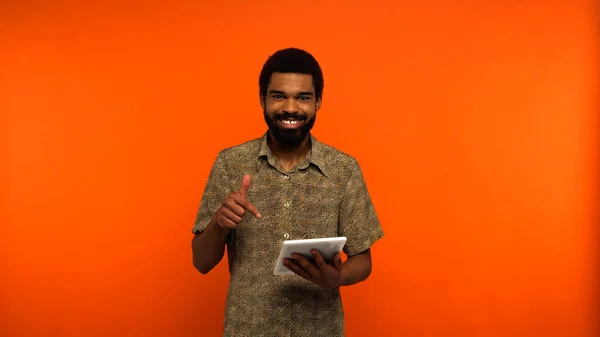 Hombre afroamericano feliz con barba apuntando con el dedo a la tableta digital sobre fondo naranja - foto de stock