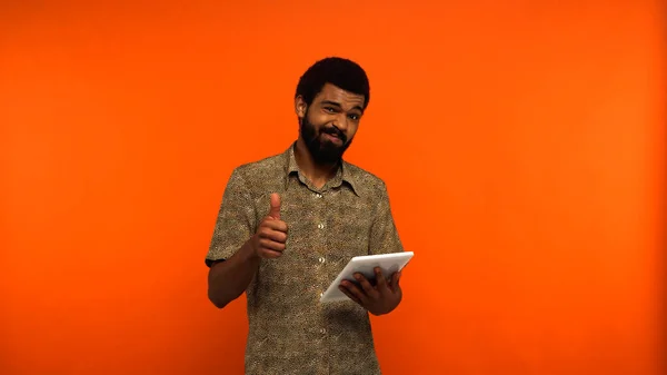 Joven afroamericano dudoso con barba sosteniendo tableta digital y mostrando como en fondo naranja - foto de stock