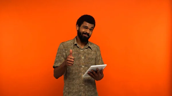 Doubtful african american young man with beard holding digital tablet and showing like on orange background — Stock Photo