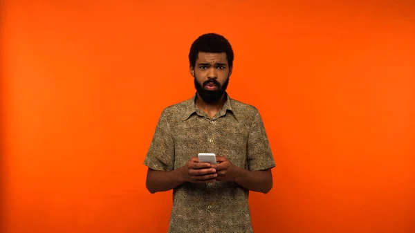 Ofendido hombre afroamericano utilizando teléfono inteligente sobre fondo naranja - foto de stock