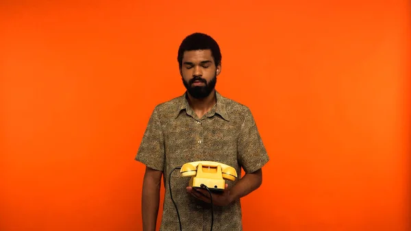 Jeune homme afro-américain aux yeux fermés tenant un téléphone rétro jaune sur fond orange — Photo de stock