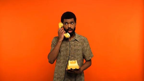 Jovem afro-americano com barba falando no telefone retro amarelo no fundo laranja — Fotografia de Stock