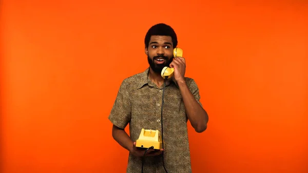 Happy african american man with beard talking on yellow retro telephone on orange background — Stock Photo