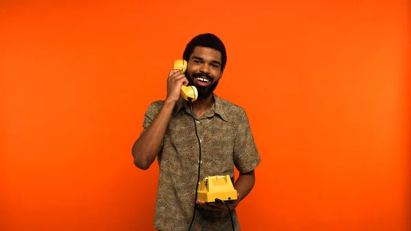 Веселый африканский американец с бородой разговаривает по ретро-телефону на оранжевом фоне — стоковое фото
