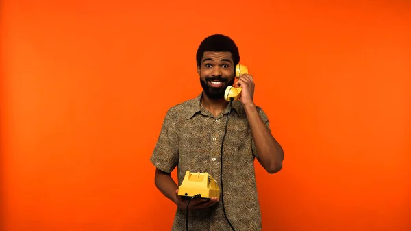 Homme afro-américain souriant avec barbe parlant sur un téléphone rétro sur fond orange — Photo de stock