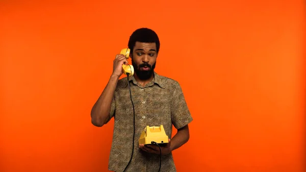 Surprised african american man with beard talking on retro telephone on orange background — Stock Photo
