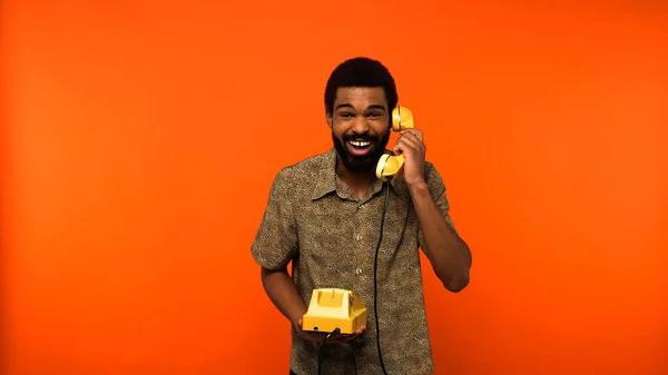 Amazed african american man with beard having conversation on retro telephone on orange background — Stock Photo