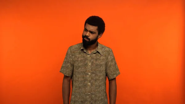Hombre afroamericano incierto con barba mirando hacia otro lado mientras está de pie sobre fondo naranja - foto de stock