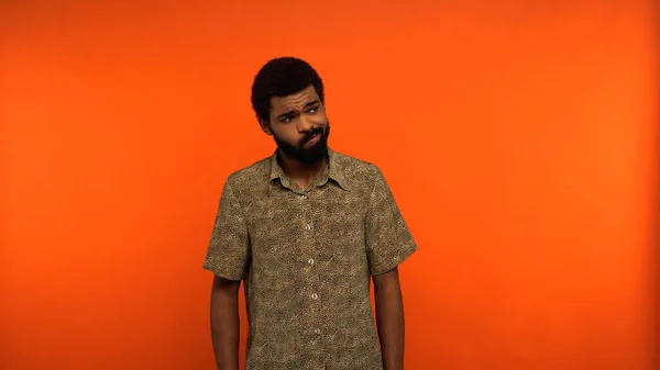 Dudoso hombre afroamericano con barba mirando hacia otro lado mientras está de pie sobre fondo naranja — Stock Photo