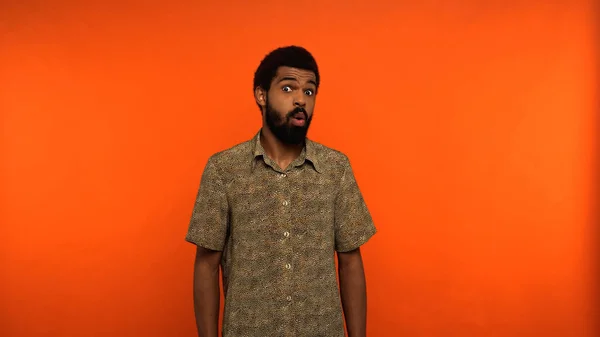 Homme afro-américain choqué avec barbe regardant la caméra sur fond orange — Photo de stock