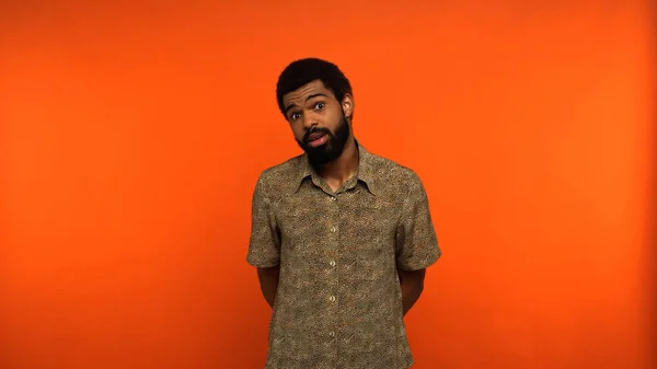 Desconcertado hombre afroamericano con barba mirando a la cámara sobre fondo naranja - foto de stock