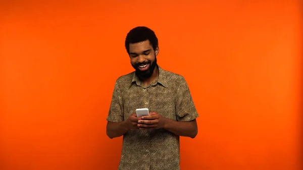 Hombre afroamericano positivo y barbudo usando teléfono inteligente sobre fondo naranja - foto de stock