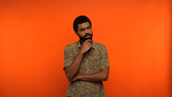 Hombre afroamericano reflexivo con barba mirando hacia otro lado mientras está de pie sobre fondo naranja - foto de stock