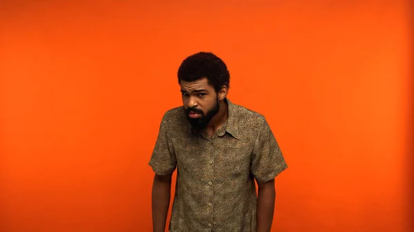 Uncertain african american man in shirt looking at camera while standing on orange background — Stock Photo