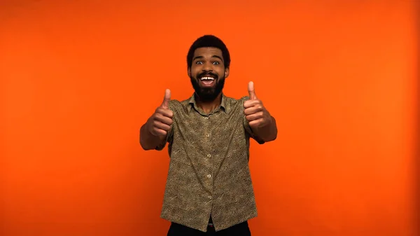 Positive african american man with opened mouth showing thumbs up on orange background — Stock Photo
