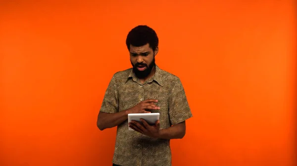 Засмучений афроамериканський молодий чоловік з бородою тримає цифровий планшет на помаранчевому фоні — стокове фото