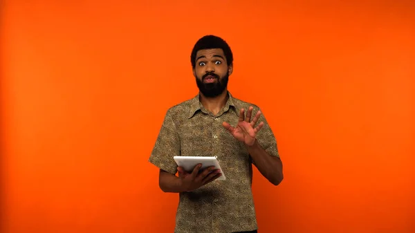 Shocked african american young man with beard holding digital tablet on orange background — Stock Photo