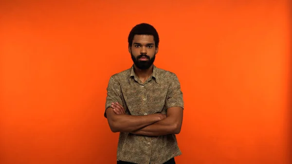 Серйозний афроамериканський молодий чоловік з бородою, що стоїть з схрещеними руками на помаранчевому фоні — стокове фото