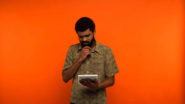 Pensativo jovem afro-americano com barba segurando tablet digital em fundo laranja — Fotografia de Stock