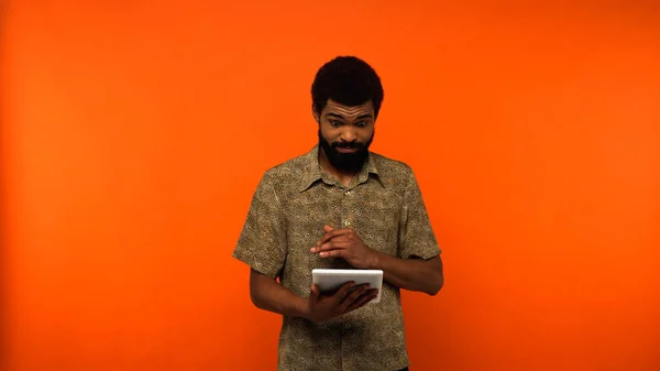 Jeune homme afro-américain confus utilisant une tablette numérique sur fond orange — Photo de stock