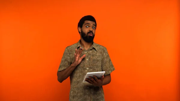 Sorprendido joven afroamericano con barba sosteniendo tableta digital mientras mira a la cámara en fondo naranja - foto de stock