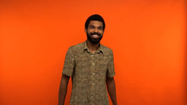 Hombre afroamericano positivo y joven con barba mirando a la cámara sobre fondo naranja - foto de stock