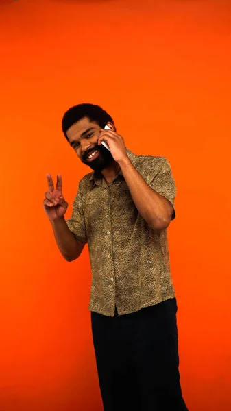 Vertical view of cheerful african american man with beard talking on smartphone and showing peace sign on orange — Stock Photo