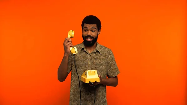 Jovem afro-americano com barba segurando telefone retro amarelo e aparelho no fundo laranja — Fotografia de Stock
