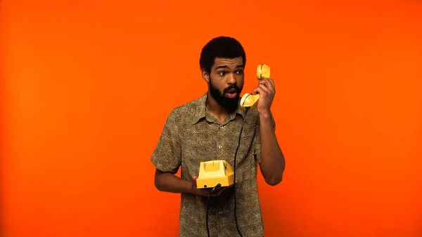 Homme afro-américain choqué avec barbe tenant téléphone rétro jaune sur fond orange — Photo de stock