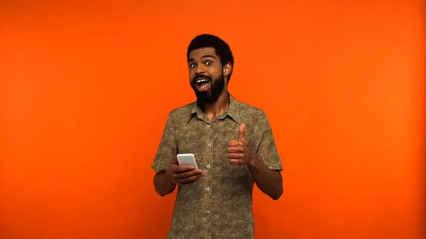 Hombre afroamericano emocionado utilizando el teléfono inteligente y mostrando como en fondo naranja - foto de stock