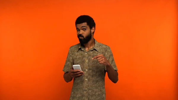 Homme afro-américain barbu pointant du doigt le smartphone sur fond orange — Photo de stock