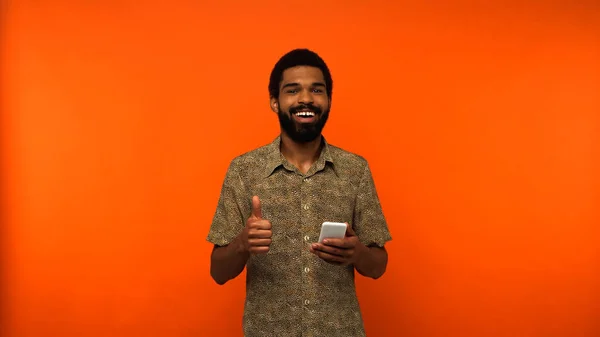 Homem americano africano positivo usando smartphone e mostrando como no fundo laranja — Fotografia de Stock