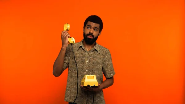 Giovane uomo africano americano con barba guardando portatile giallo del telefono retrò su sfondo arancione — Foto stock