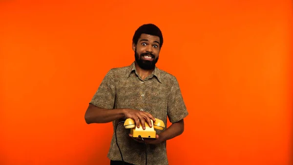 Confuso homem americano africano com barba segurando telefone retro amarelo no fundo laranja — Fotografia de Stock