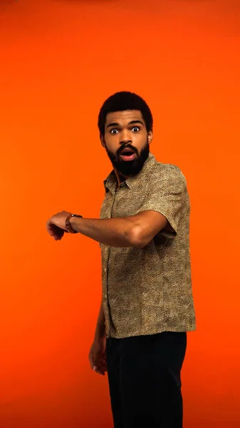 Visão vertical do jovem afro-americano chocado com relógio de pulso olhando para a câmera em laranja — Fotografia de Stock