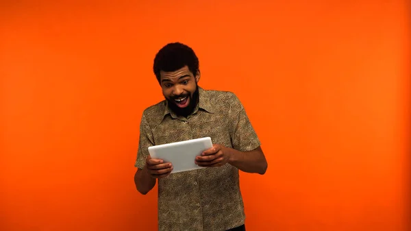 Espantado jovem afro-americano com barba usando tablet digital em fundo laranja — Fotografia de Stock