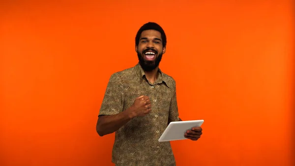Excité jeune homme afro-américain avec barbe tenant tablette numérique sur fond orange — Photo de stock