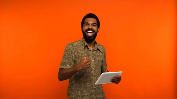 Joven afroamericano emocionado con barba sosteniendo tableta digital sobre fondo naranja - foto de stock
