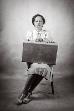 Geçmişe dönük giyinmiş genç bir kadın, bir sandalyede oturuyor, dizlerinde bir bavulla. Stüdyo siyah beyaz çekim