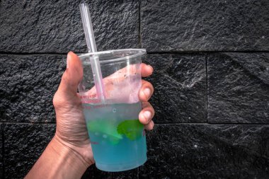 Elinde taze mavi mojito, şeffaf plastik bardakta, arka planda siyah taşlarla birlikte.