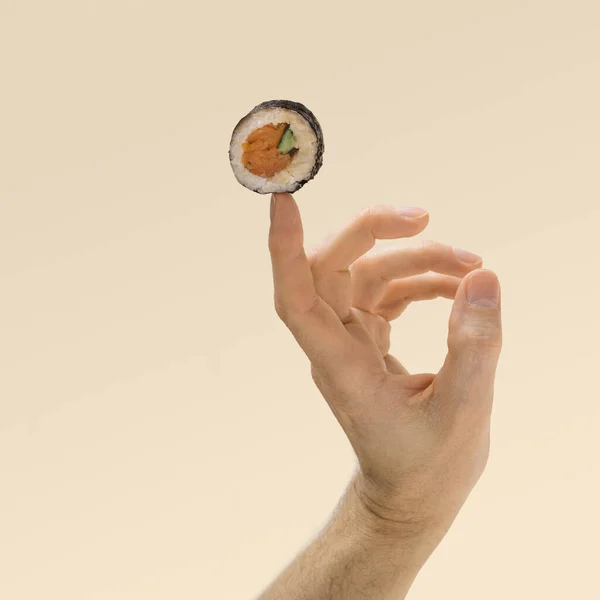 Sushi minimal concept. Man hand balance with one piece rice, salmon and cucumber roll in nori dried edible seaweed on index finger. Awesome beige background