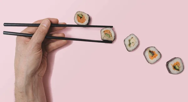 Flat lay sushi vegetarian concept. Man hand holding black chopsticks grabbing one piece, salmon fish, rice, cucumber and nori seaweed between many others different sushi rolls. Pastel pink background