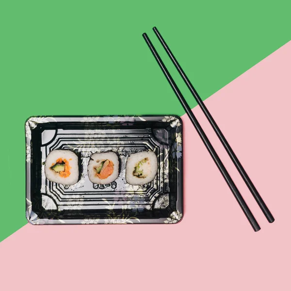 Three sushi with salmon, squid, tuna fish, rice, cucumber carrots  and nori seaweed rolls in a rectangular disposable black plate on diagonal pastel pink and green background with chopsticks. Takeaway flat lay sushi concept.