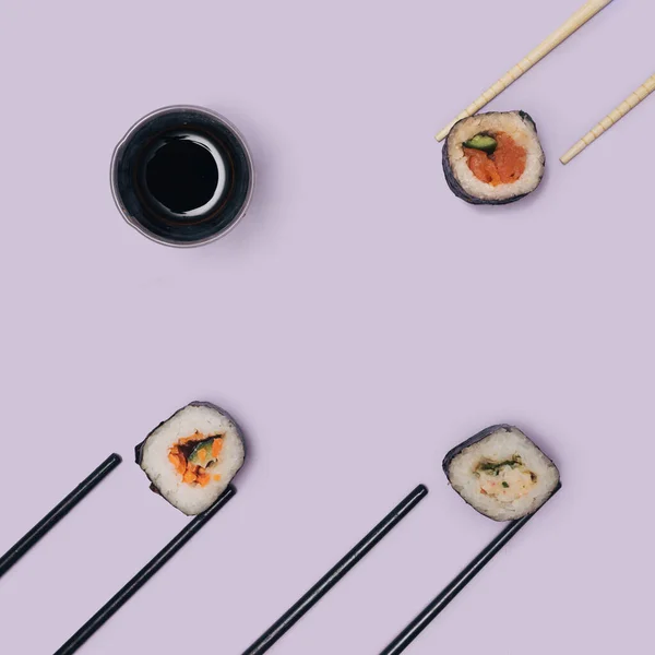 Flat lay sushi concept. Three sushi with salmon, squid, tuna fish, rice, cucumber carrots  and nori seaweed rolls touching chopsticks and a little glass with soy sauce. Very peri background
