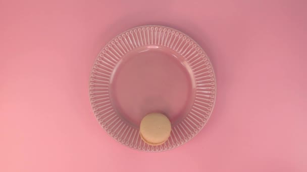 A womans hand puts pink cup of coffee with milk cream on the pink plate with macaroon on the pink table. Top view to the table. Romantic dessert. — стоковое видео