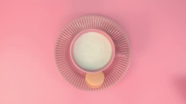 Une main de femme prend une tasse de café rose avec de la crème de lait sur l'assiette rose avec un macaron sur la table rose. Vue du dessus de la table. Dessert romantique. — Video