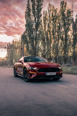 Güzel günbatımının arka planında kırmızı renkli Amerikan kaslı arabası Ford Mustang. Kherson, Ukrayna - Mayıs 2021.