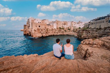 Polignano a Mare, İtalya - Ağustos 2021: Kayalıklarda oturan genç çift yazın Polignano a Mare köyünü izliyor