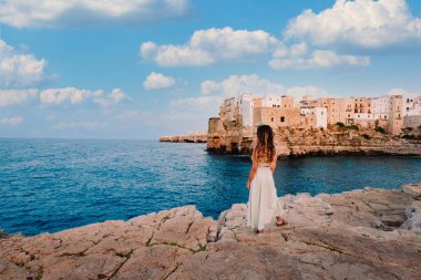 Polignano a Mare, İtalya - Ağustos 2021: Polignano a Mare köyünün önündeki genç kız, gökyüzü beyaz bulutlu