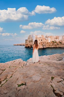 Polignano a Mare, İtalya - Ağustos 2021: Polignano a Mare köyünün önündeki genç kız, gökyüzü beyaz bulutlu, dikey